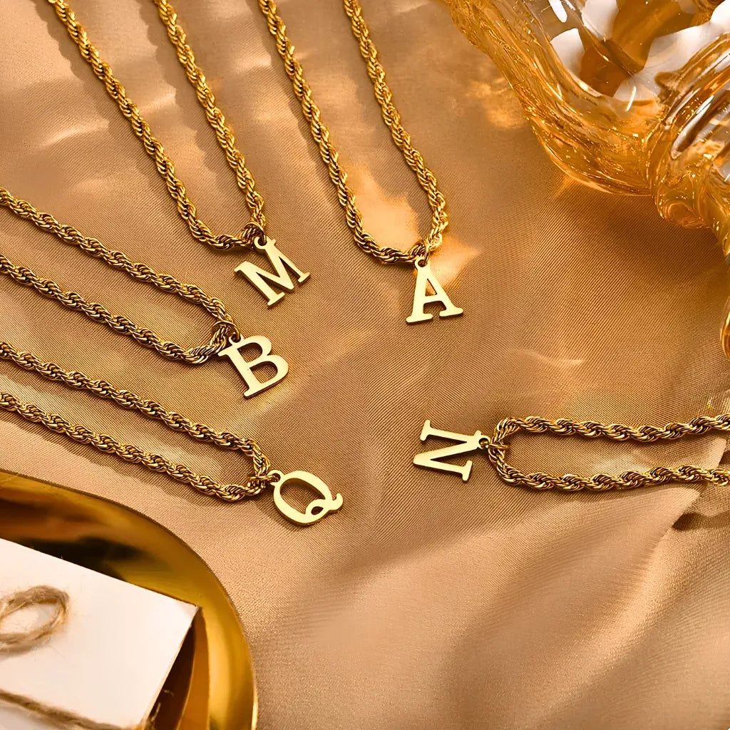 Colares dourados com pingentes de letras M, A, B, Q e N sobre tecido bege, acessórios femininos