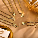 Colares dourados com pingentes de letras M, A, B, Q e N sobre tecido bege, acessórios femininos