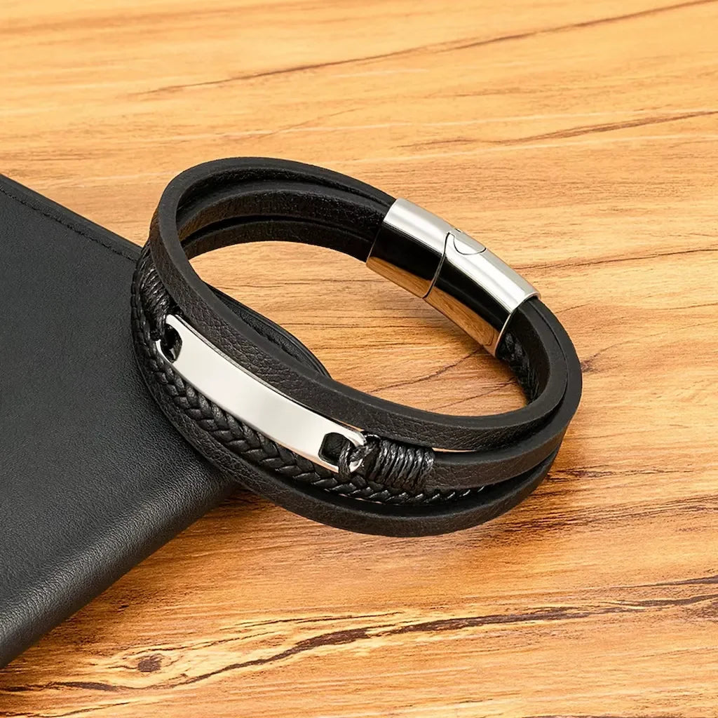 Pulseira masculina preta de couro com detalhe trançado e placa metálica prata sobre superfície de madeira